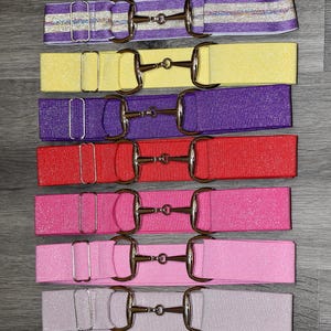 May include: A collection of colorful, glittery elastic belts with silver-colored metal bit buckles. The belts are in various shades, including purple, yellow, red, and pink. The belts are approximately 1.5 inches wide.
