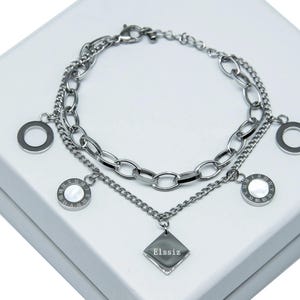 Könnte beinhalten: Ein silberfarbenes Armband mit mehreren Ketten und Anhängern. Das Armband verfügt über eine Kombination aus Kettenstilen, darunter ovale und Curb-Glieder. Zu den Anhängern gehören offene Kreise, römische Ziffernscheiben und ein rautenförmiger Anhänger mit dem Text "Elssiz".