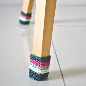 Könnte beinhalten: Nahaufnahme eines Holzstuhlbeins mit einer gestrickten Socke in den Farbtönen Türkis, Rosa, Weiß und Schwarz. Die gestrickte Socke bedeckt die Unterseite des Stuhlbeins und schützt den Boden vor Kratzern.