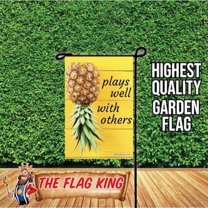 May include: A yellow garden flag with a pineapple illustration and the text "plays well with others". The flag is attached to a black metal pole. The flag is on a green background.
