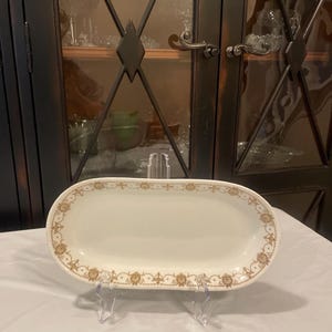 May include: A white oval serving dish with a gold floral pattern around the edge. The dish is on a white surface.