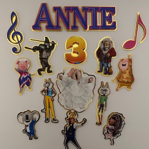 May include: Cake toppers featuring characters from the animated film "Sing". The toppers include the name "ANNIE" in blue letters with a gold outline, the number "3", musical notes, and various character cutouts.