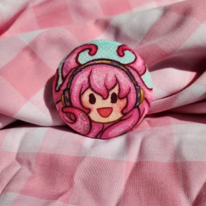 May include: A round, pink button featuring a cartoon character with pink hair, headphones, and a smiling face. The button has a light blue background with a polka dot pattern. The button is set against a pink and white checkered fabric.