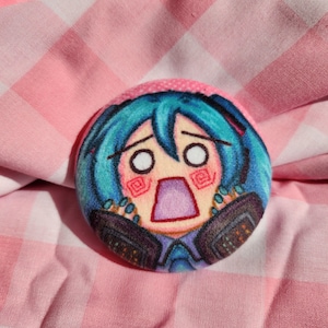 May include: A round button featuring a cartoon character with teal hair and a surprised expression. The character's face is pink and white, with large, wide eyes and an open mouth. The button is set against a pink and white checkered background.