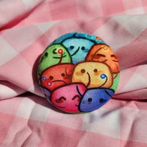 May include: A round, colorful pin featuring a collection of smiling cartoon faces in various colors, including green, blue, orange, and pink. The pin is set against a pink and white checkered fabric background.