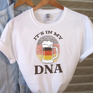 May include: White t-shirt with a brown graphic of a beer mug with a German flag inside a fingerprint. The text "IT'S IN MY DNA" is printed below the graphic.