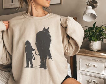 May include: A beige sweatshirt with a black silhouette of a person leading a horse. The sweatshirt has a relaxed fit and a crew neck.