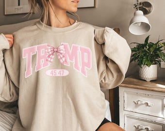 May include: A beige sweatshirt with a pink graphic of the word "TRUMP" with a pink and white gingham bow. The numbers "45 47" are printed below the graphic.
