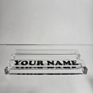 Puede incluir: Placa de escritorio de acr&iacute;lico transparente con el texto "YOUR NAME" en letras negras en negrita. La placa tiene forma rectangular y es transparente. Una cubierta de acr&iacute;lico transparente se encuentra encima.