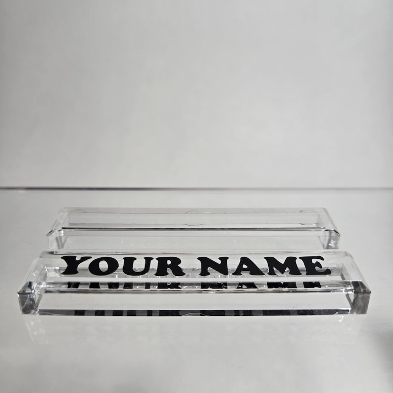 Puede incluir: Placa de escritorio de acr&iacute;lico transparente con el texto "YOUR NAME" en letras negras en negrita. La placa tiene forma rectangular y es transparente. Una cubierta de acr&iacute;lico transparente se encuentra encima.