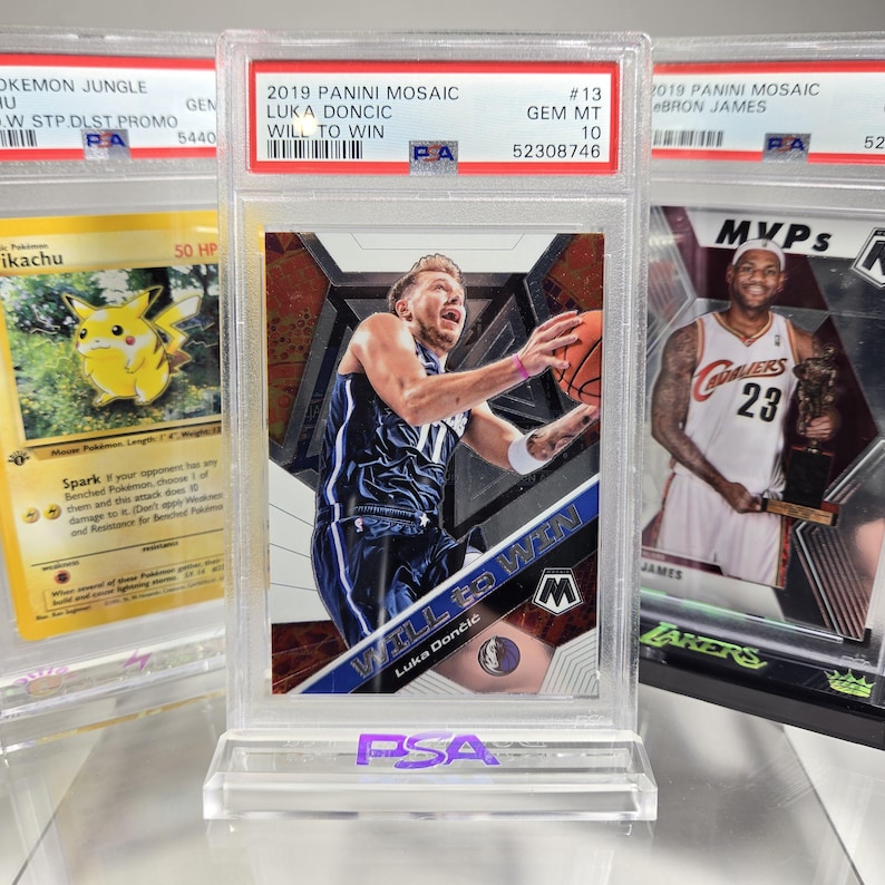 Puede incluir: Una tarjeta de baloncesto de Luka Dončić de 2019 Panini Mosaic, calificada, con el texto "WILL TO WIN". La tarjeta est&aacute; en un soporte protector y se muestra con otras tarjetas coleccionables, incluyendo una tarjeta Pok&eacute;mon y una tarjeta LeBron James.