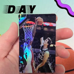 May include: A basketball card featuring a player in a white and red jersey attempting a dunk, with a player in a black and silver jersey defending. The card has the words "GAME DAY" in black above the image. A basketball is visible in the background.