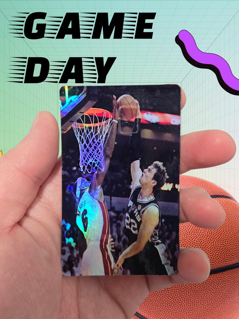 May include: A basketball card featuring a player in a white and red jersey attempting a dunk, with a player in a black and silver jersey defending. The card has the words "GAME DAY" in black above the image. A basketball is visible in the background.