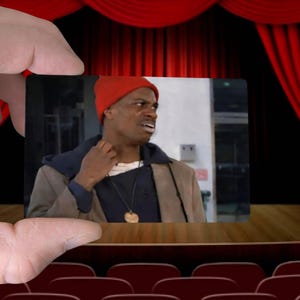 May include: A small image of a man wearing a red beanie, a brown coat, and a necklace is held in front of a theater stage with red curtains. The man appears to be looking off to the side with a concerned expression.