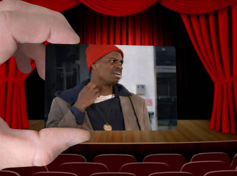 May include: A small image of a man wearing a red beanie, a brown coat, and a necklace is held in front of a theater stage with red curtains. The man appears to be looking off to the side with a concerned expression.