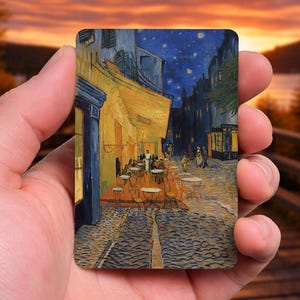 May include: A rectangular art print of Van Gogh's "Cafe Terrace at Night" held in a hand. The painting depicts a nighttime street scene with a cafe, tables, and figures under a starry sky. The print has a warm, inviting color palette.