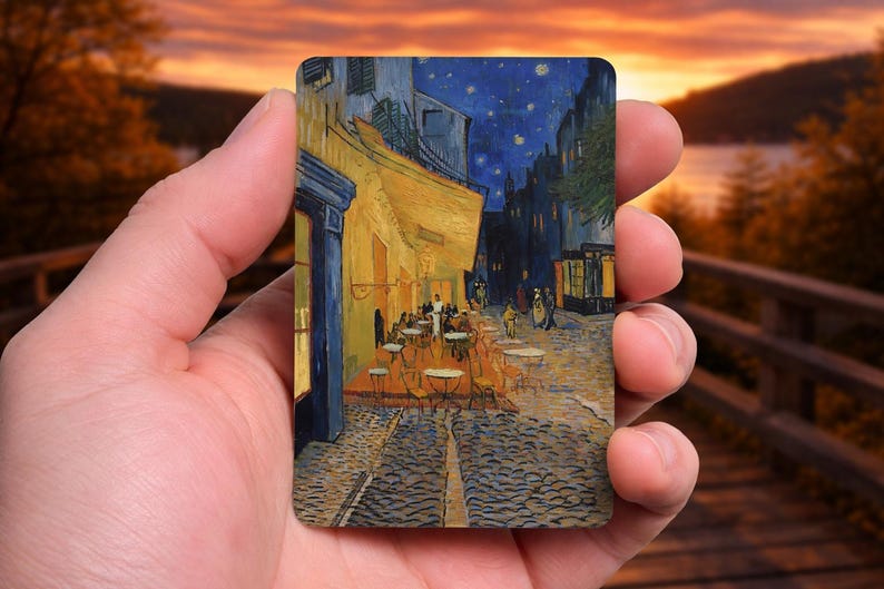 May include: A rectangular art print of Van Gogh's "Cafe Terrace at Night" held in a hand. The painting depicts a nighttime street scene with a cafe, tables, and figures under a starry sky. The print has a warm, inviting color palette.