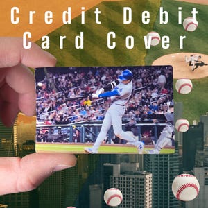 May include: A custom credit/debit card cover featuring a baseball player mid-swing, set against a stadium crowd. The background includes baseballs and cityscapes, with the words "Custom Credit Debit Card Cover" in white.