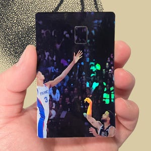 May include: A rectangular black card featuring a basketball player in a blue, white, and red uniform, reaching for the ball. The card has a glossy finish and a basketball design in the background.