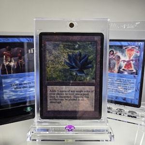 May include: A black lotus magic card in a clear plastic protective case. The card is from the Alpha set and features a black lotus flower on a dark green background. The text on the card reads "Black Lotus" and "Adds 3 mana of any single color of your choice to your mana pool, then is discarded. Tapping this artifact can be played as an interrupt."