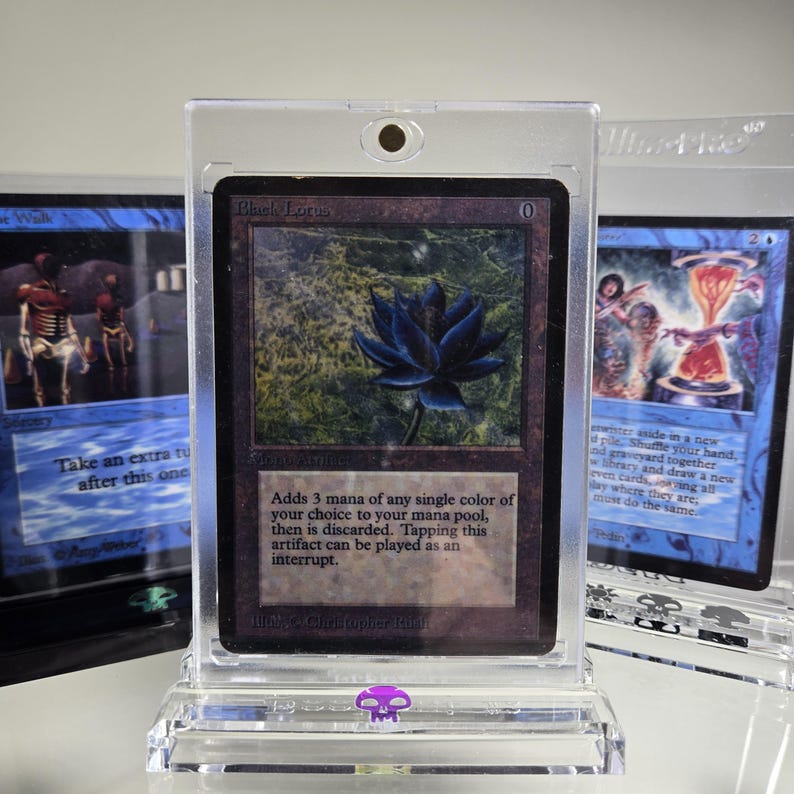 May include: A black lotus magic card in a clear plastic protective case. The card is from the Alpha set and features a black lotus flower on a dark green background. The text on the card reads "Black Lotus" and "Adds 3 mana of any single color of your choice to your mana pool, then is discarded. Tapping this artifact can be played as an interrupt."