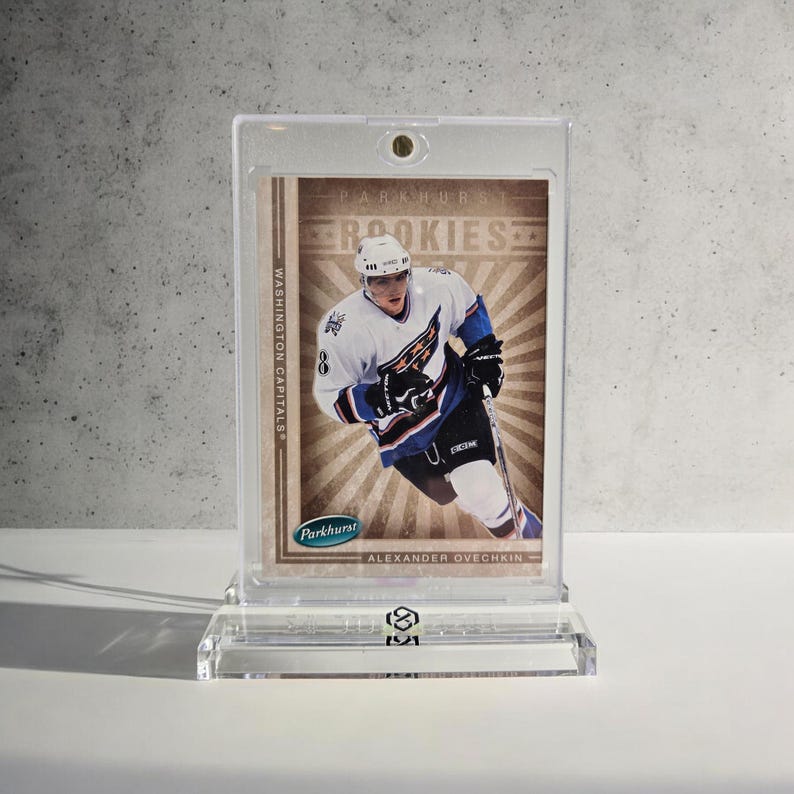 Puede incluir: Una tarjeta de hockey con Alexander Ovechkin con el uniforme de los Washington Capitals, exhibida en un soporte y expositor de acr&iacute;lico transparente. La tarjeta tiene el texto "Parkhurst Rookies" y "Washington Capitals".