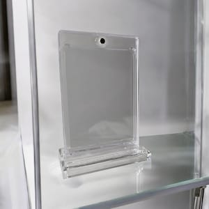 May include: A clear acrylic sign holder with a rectangular design. The sign holder has a base and a slot to hold a sign. A small hole is at the top of the sign holder.
