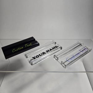 Puede incluir: Tres placas de escritorio rectangulares. Una es negra con "Custom Text" en verde. Dos son de acr&iacute;lico transparente con "YOUR NAME" y "Custom Text" en negro y azul, respectivamente. Todas est&aacute;n sobre una estanter&iacute;a transparente.
