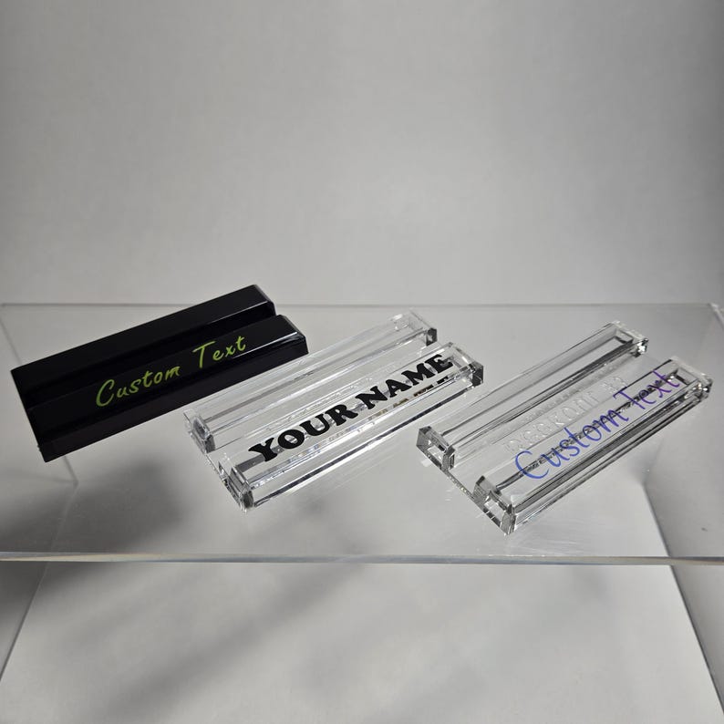 Puede incluir: Tres placas de escritorio rectangulares. Una es negra con "Custom Text" en verde. Dos son de acr&iacute;lico transparente con "YOUR NAME" y "Custom Text" en negro y azul, respectivamente. Todas est&aacute;n sobre una estanter&iacute;a transparente.