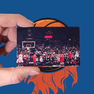 May include: A rectangular card featuring a basketball game scene, held against a blue background with an orange basketball graphic. The card shows players in red and white uniforms, with a crowd in the background. The text "PLAYOFF" is visible.