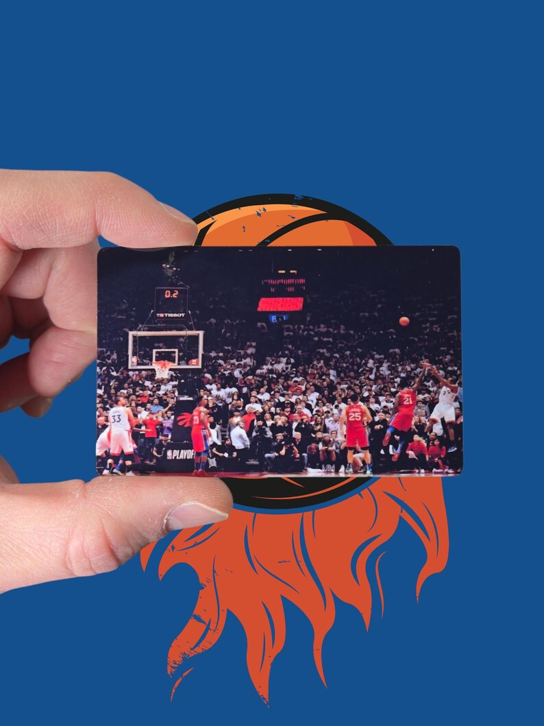 May include: A rectangular card featuring a basketball game scene, held against a blue background with an orange basketball graphic. The card shows players in red and white uniforms, with a crowd in the background. The text "PLAYOFF" is visible.
