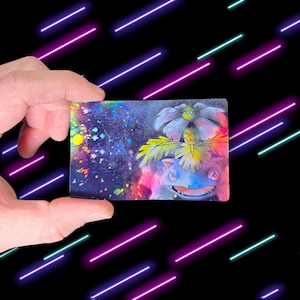 May include: A rectangular card featuring a colorful illustration of a blue and green cartoon character with a flower on its back. The background has a galaxy-like design with splashes of color. The card is held against a black background with neon lights.