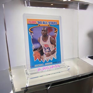 Puede incluir: Una tarjeta de baloncesto coleccionable con Michael Jordan con una camiseta de la NBA blanca y roja con el n&uacute;mero 23. La tarjeta es de la colecci&oacute;n Fleer '90 All-Stars y se muestra en un soporte acr&iacute;lico transparente.