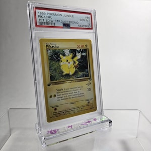 Puede incluir: Una carta Pok&eacute;mon de Pikachu del set Jungle de 1999, calificada. La carta est&aacute; dentro de un soporte protector transparente con una etiqueta PSA. La carta tiene un fondo amarillo con una ilustraci&oacute;n de Pikachu y texto.