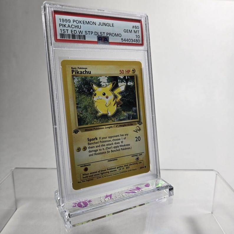 Puede incluir: Una carta Pok&eacute;mon de Pikachu del set Jungle de 1999, calificada. La carta est&aacute; dentro de un soporte protector transparente con una etiqueta PSA. La carta tiene un fondo amarillo con una ilustraci&oacute;n de Pikachu y texto.