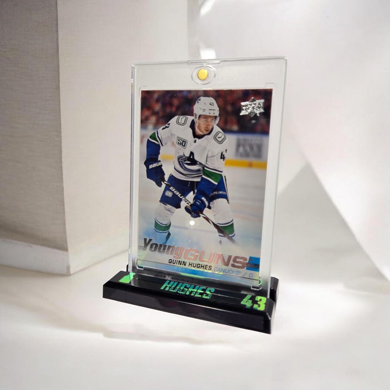 Puede incluir: Una tarjeta de hockey de Quinn Hughes en una vitrina acr&iacute;lica transparente. La tarjeta presenta a Hughes con el uniforme de los Canucks de Vancouver. La vitrina tiene una base negra con el nombre "HUGHES" y el n&uacute;mero "43" en verde.