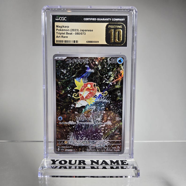 Puede incluir: Una carta de Pok&eacute;mon Magikarp calificada en una funda protectora transparente. La carta presenta una colorida ilustraci&oacute;n de una criatura con forma de pez con el texto "Magikarp" y "Triplet Beat - 080/073". La funda est&aacute; etiquetada como "PRISTINE 10".