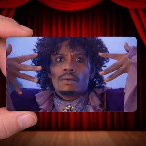 May include: A rectangular card featuring a person with curly hair, wearing a purple outfit with a ruffled collar. The person is making a hand gesture. The background is a red curtain.