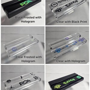 May include: Six different acrylic display stands for trading cards. The stands are clear, frosted, and black with holographic and black printed designs. The stands feature the text "PSA" and "Pokemon".