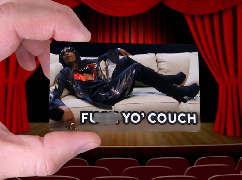 May include: A rectangular card held by a hand, featuring an image of a person reclining on a couch. The person is wearing black clothing with silver accents. The card has the text "F*** Yo' Couch". The background includes red curtains.