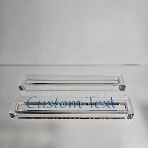 Puede incluir: Dos expositores de acr&iacute;lico transparente, uno apilado sobre el otro. El soporte inferior tiene las palabras "Custom Text" en azul. El soporte superior tiene las palabras "Deekord #3" en azul.