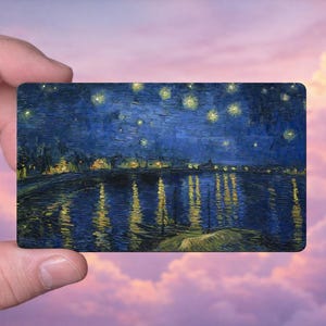 May include: A rectangular card featuring a painting of a night scene with a river reflecting lights and stars. The artwork is dominated by dark blues and yellows, reminiscent of Van Gogh's style. The card is held against a backdrop of a pink and purple sky.