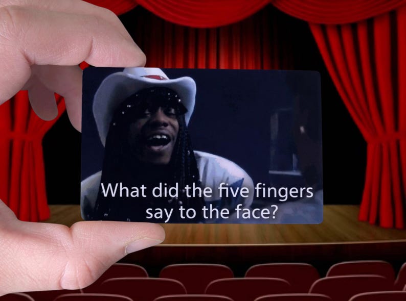 May include: A rectangular card held by a hand displays a scene with a person wearing a cowboy hat. The text on the card reads, "What did the five fingers say to the face?" The background features a stage with red curtains and theater seating.