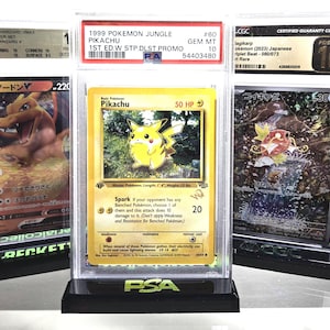 May include: Three graded Pokémon trading cards in protective cases. The center card features Pikachu from the 1999 Jungle set. The other cards feature Charizard and Magikarp. The cards are displayed on black stands.