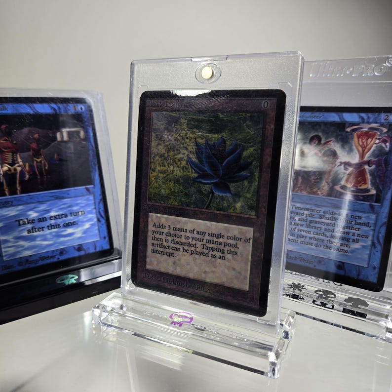 May include: Three Magic: The Gathering cards in protective plastic cases. The card in the center is a black lotus card with a black flower on a green background. The text on the card reads "Adds 3 mana of any single color of your choice to your mana pool, then is discarded. Tapping this artifact can be played as an interrupt."