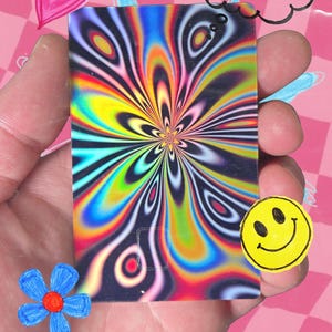 Pu&ograve; includere: Una carta rettangolare con un vivace design psichedelico nei toni del blu, giallo, verde e rosso &egrave; tenuta in mano. La carta presenta un motivo a spirale a forma di fiore. Un adesivo giallo con la faccina sorridente e altri scarabocchi circondano la carta.