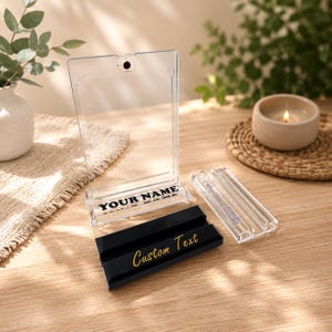 Puede incluir: Un expositor de acr&iacute;lico transparente con los textos "YOUR NAME" y "Custom Text" en negro y dorado. El expositor est&aacute; sobre una superficie de madera, con una vela y vegetaci&oacute;n en el fondo.
