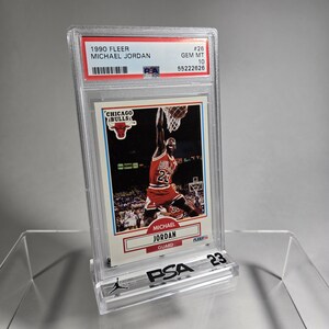 Puede incluir: Una tarjeta de baloncesto de Michael Jordan de 1990 Fleer, calificada GEM MT 10 por PSA, en un estuche protector transparente. La tarjeta muestra a Michael Jordan con el uniforme rojo de los Chicago Bulls, en el aire, a punto de encestar.