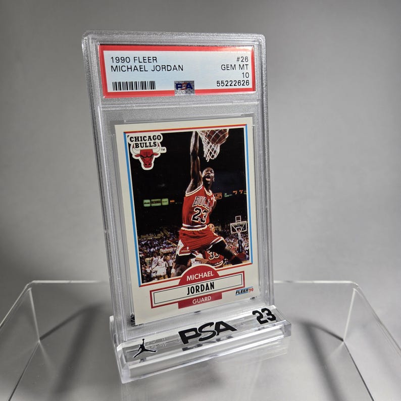 Puede incluir: Una tarjeta de baloncesto de Michael Jordan de 1990 Fleer, calificada GEM MT 10 por PSA, en un estuche protector transparente. La tarjeta muestra a Michael Jordan con el uniforme rojo de los Chicago Bulls, en el aire, a punto de encestar.