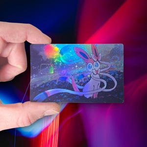 May include: A holographic trading card featuring a Sylveon character. The card has a colorful, iridescent design with a pink, white, and blue color scheme. The card is rectangular and held by a hand.